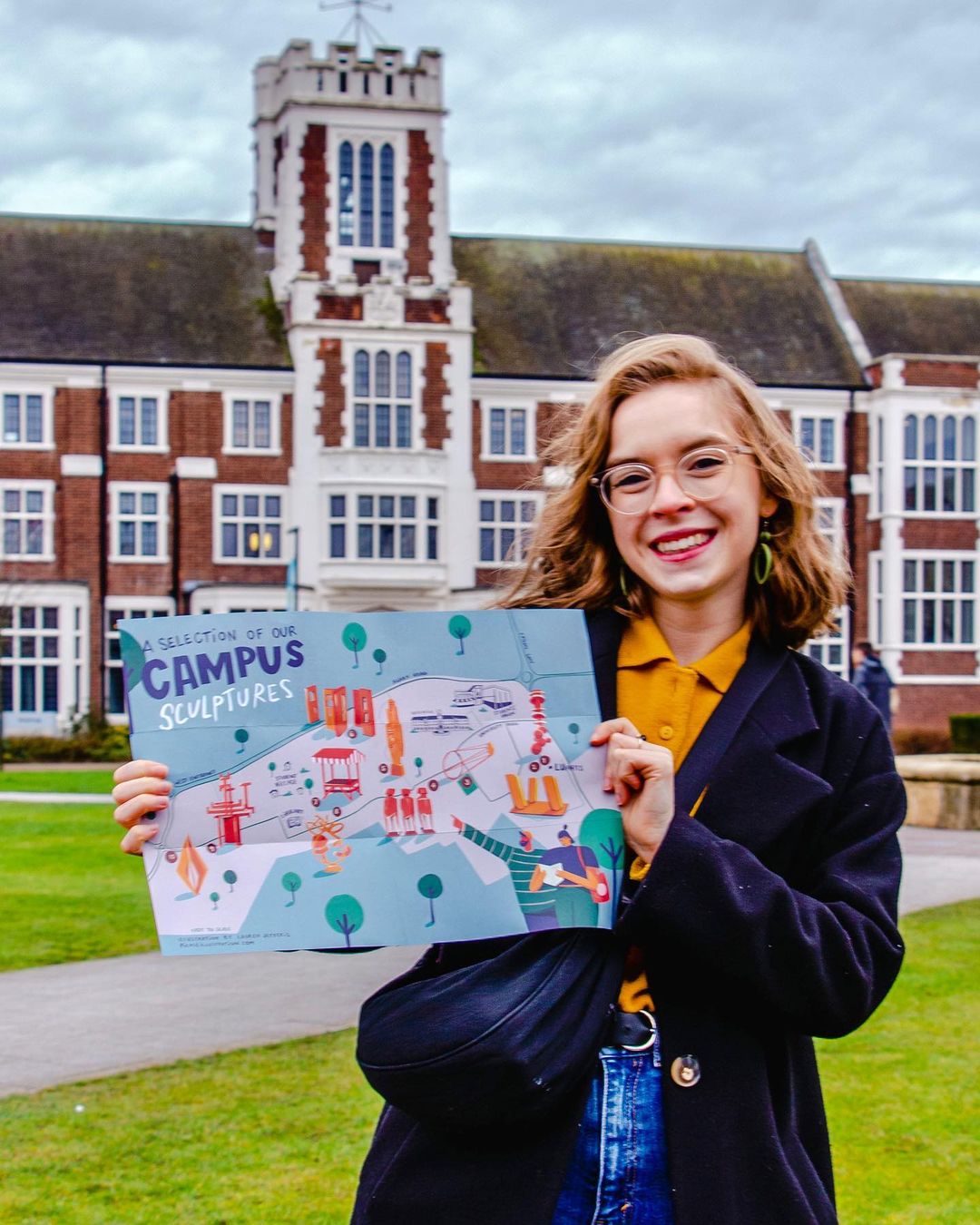 A white woman with short blonde hair and glasses smiles in front of the Hazlerigg building holding a blue and green illustrated map